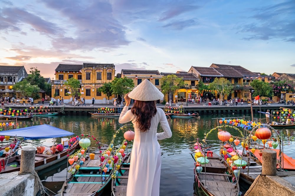 A woman in a traditional Vietnamese dress stands gracefully on a river dock, surrounded by serene water and nature, at Hoi An ancient town.